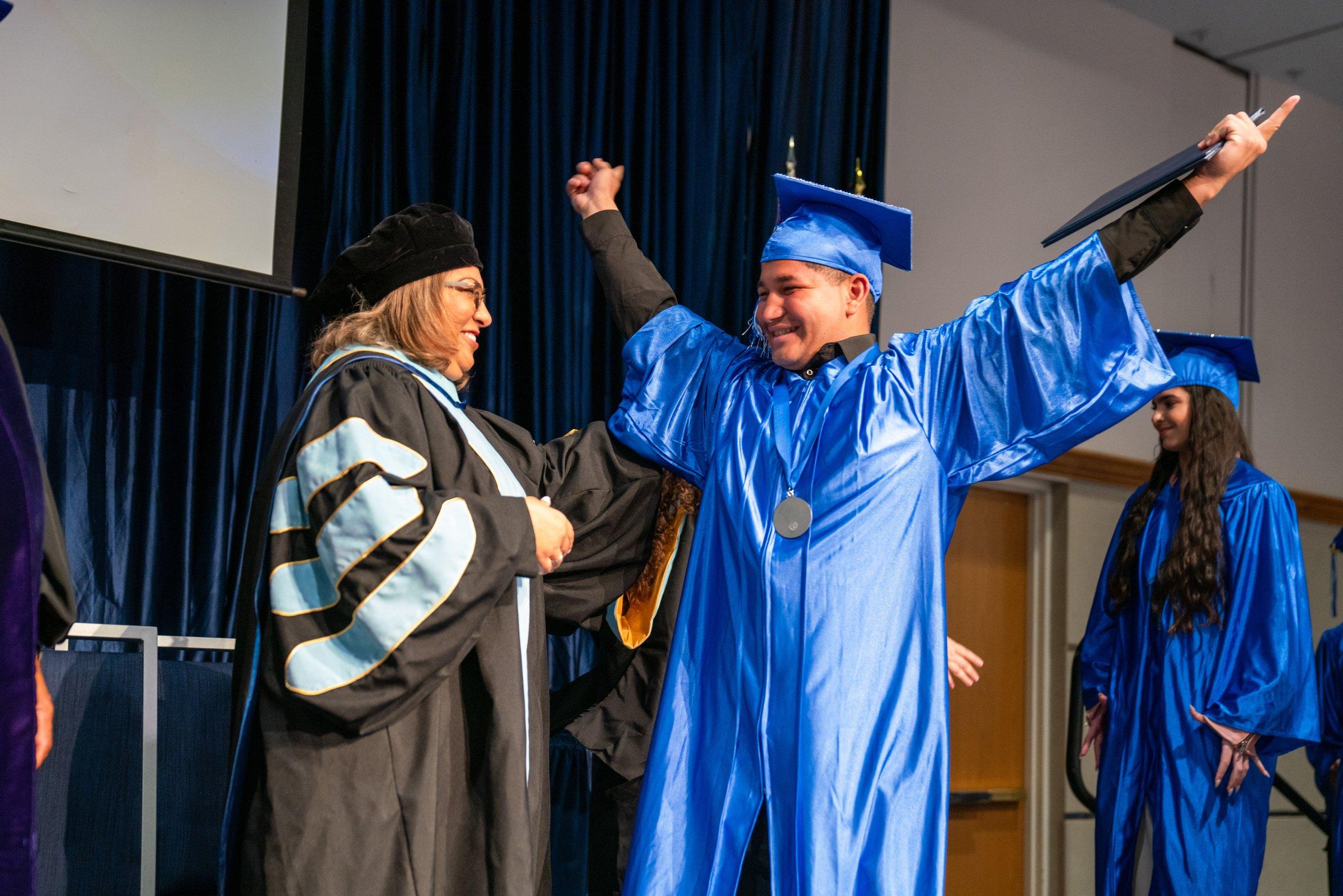 A graduate raising both arms in celebration on stage beside a smiling administrator